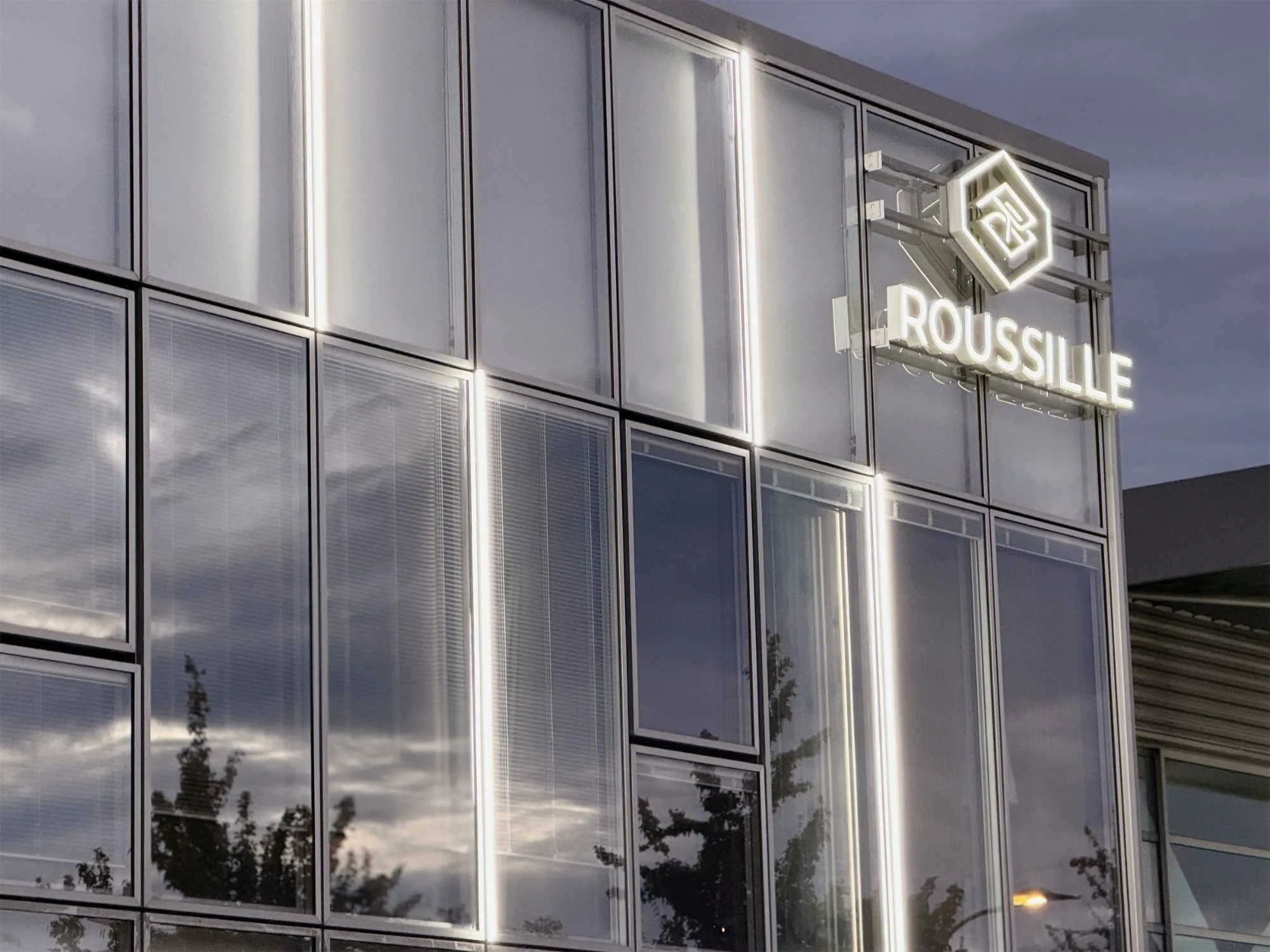 Façade vitrée du siège de Roussille Assurances à Pau, enseigne et logo lumineux à la tombée de la nuit